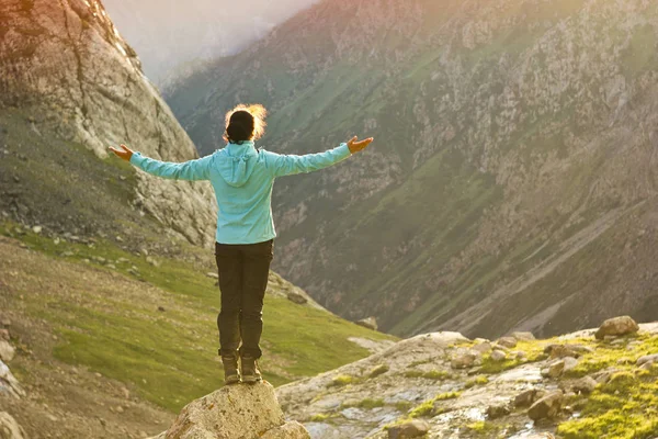 Girl in blue jacket standing on stone with hands-up above mountais at sunset — Stock Photo, Image