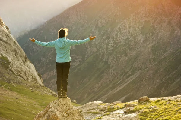 Niña en chaqueta azul de pie sobre piedra con las manos arriba por encima de mountais al atardecer — Foto de Stock