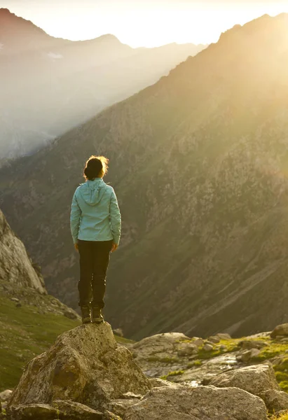 Girl in blue jacket standing on stone with hands-up above mountais at sunset — Stock Photo, Image