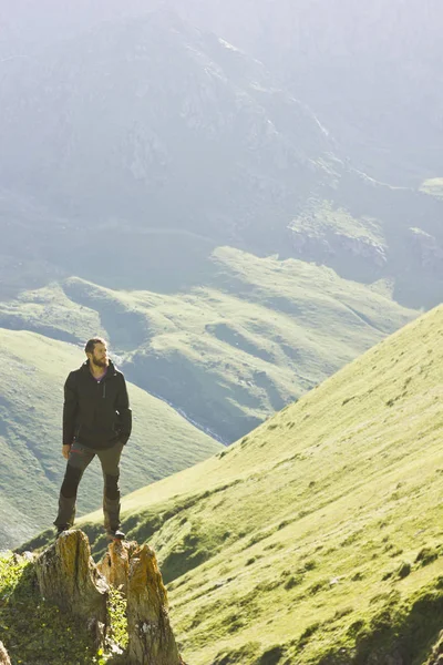 Man in black jacket standing on stone with hands-up above mountais at sunset — Stock Photo, Image