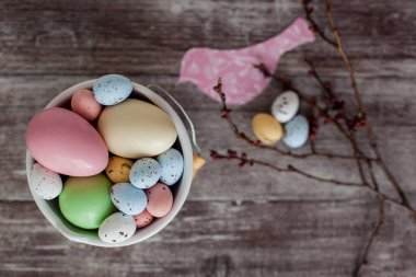 Yumurtalı Paskalya kompozisyonu, tomurcuklu bir ağaç filizi ve koyu ahşap arka planda dekoratif bir kuş. Beyaz seramik kovada farklı boyutlarda pastel renkli Paskalya yumurtaları.