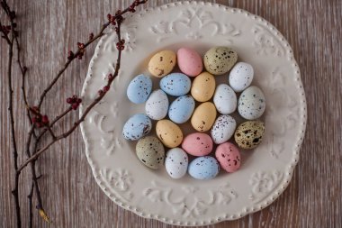 Tahta bir masanın üzerinde açık bir tabakta, çok renkli Paskalya yumurtalarının yanında henüz çiçek açmamış kayısı tomurcukları olan dallar var. Üst Görünüm