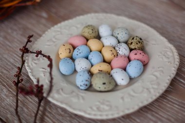 Tahta bir masanın üzerinde açık bir tabakta çok renkli Paskalya yumurtaları, hemen yanında henüz çiçek açmamış kayısı tomurcukları olan dallar var..