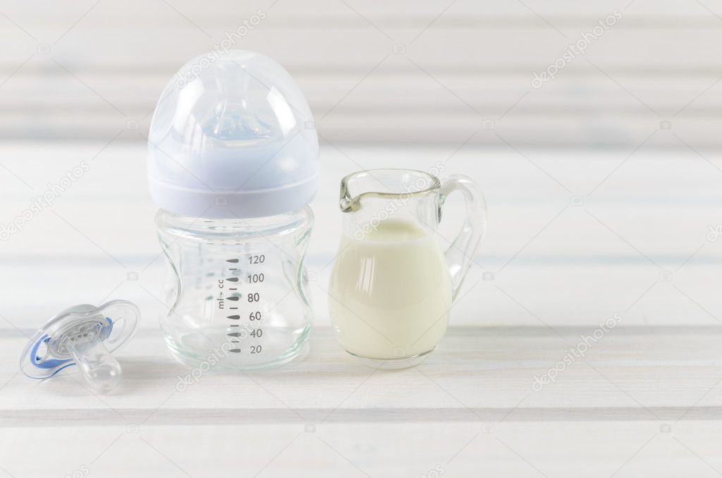 Glass baby bottle, milk jug and baby pacifier on white wooden ba