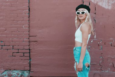 Güneş gözlüklü ve siyah şapkalı bir duvarın yanında duran genç kadın.