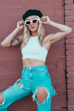 Güneş gözlüklü ve siyah şapkalı bir duvarın yanında duran genç kadın.