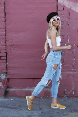Güneş gözlüklü ve siyah şapkalı bir duvarın yanında duran genç kadın.