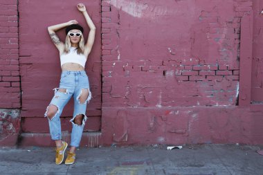 Güneş gözlüklü ve siyah şapkalı bir duvarın yanında duran genç kadın.