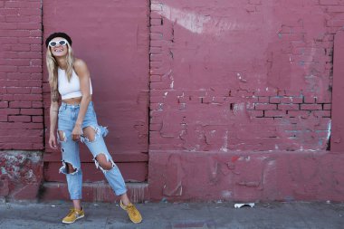 Güneş gözlüklü ve siyah şapkalı bir duvarın yanında duran genç kadın.