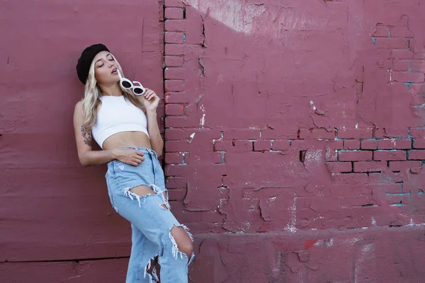 Mujer joven de pie junto a una pared con gafas de sol y sombrero negro —  Fotos de Stock