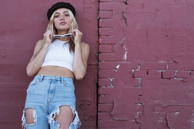 Güneş gözlüklü ve siyah şapkalı bir duvarın yanında duran genç kadın.