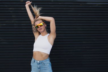 Güneş gözlüklü, siyah bir duvarın yanında duran genç bir kadın.