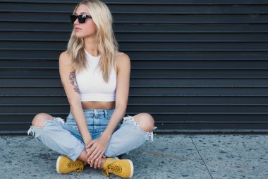 Güneş gözlüklü, siyah bir duvarda oturan genç bir kadın.