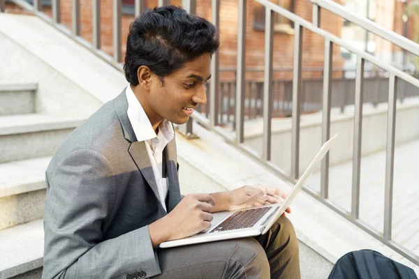 Young handsome Indian man is sitting with a laptop, smiling and typing