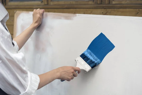 Female hand painting a white sheet of plywood by blue paint