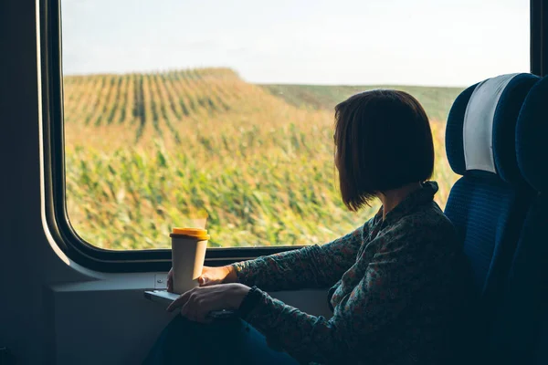 A woman sitting by the window of a commuter train with a laptop and coffee cup