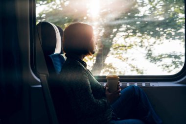 Bir kadın, bir banliyö treninin penceresinde dizüstü bilgisayarı ve kahve fincanıyla oturuyor.