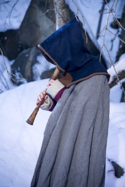 Guy in a medieval costume with a hood playing on a pipe, in the winter clipart