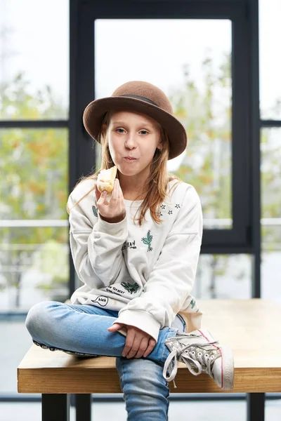 Ragazza divertente in un grande cappello mangia una torta dolce, si siede su un tavolo nella stanza — Foto Stock