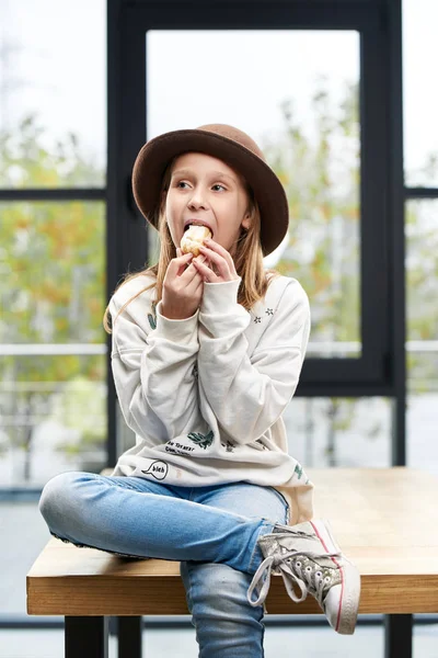 Забавная девушка в большой шляпе ест сладкий торт, сидит на столе в комнате — стоковое фото