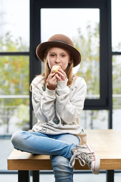 Ragazza divertente in un grande cappello mangia una torta dolce, si siede su un tavolo nella stanza — Foto Stock