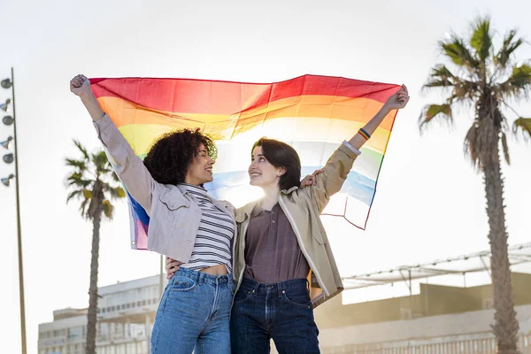 Giovane coppia lesbica sorridente sotto una bandiera arcobaleno — Foto Stock
