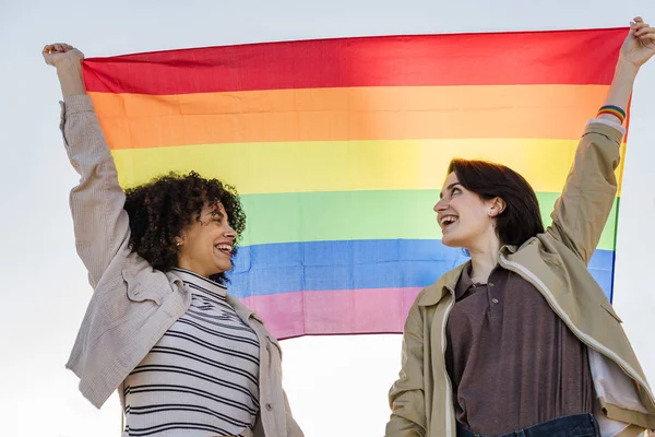 Casal de mulheres sorridentes levantando a bandeira do arco-íris — Fotografia de Stock