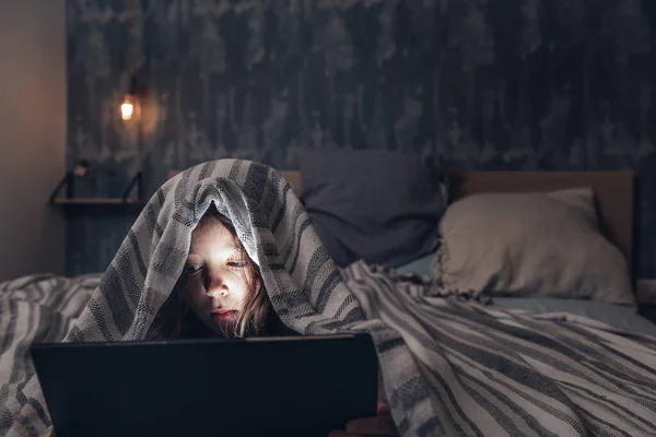 little girl lying in bed covered up to the head with a quilt watching a movie on a tablet in the darkness with the face illuminated by the screen, child and technology concept, copy space for text