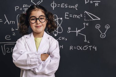 Laboratuvar önlüklü neşeli küçük kız fen öğrencisi okul karatahtasında gülümsüyor. El çizimleri, bilimsel formül kalıpları, okula geri dönüş ve başarılı kadın kariyer kavramı.