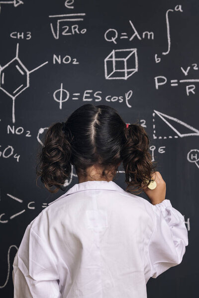 rear view of a little girl science student in lab coat writing on school blackboard with hand drawings science formula pattern, back to school and successful female career concept, vertical photo