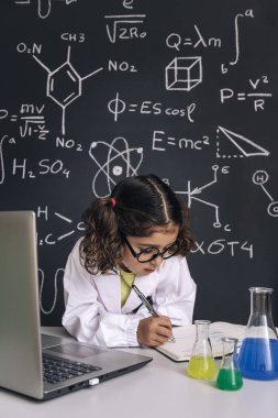 Gözlüklü, bilgisayarla çalışan ve notlar alan bilim formüllü okul karatahtası geçmişi olan okula geri dönen ve başarılı kadın kariyer konsepti, dikey fotoğraf çeken küçük bir bilim adamı kız.