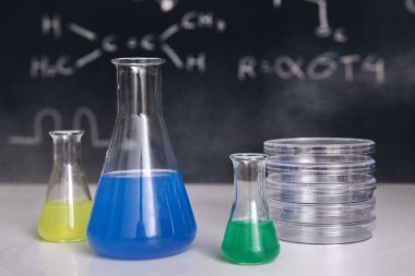 İçinde renkli kimyasallar, enstrümanlar olan mataralarla dolu bir sınıf masası ve arka planda fen bilgisi öğreten çizilmiş bir karatahta, kimyasal bilimler eğitim kavramı