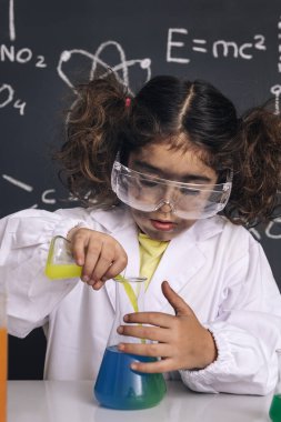 Gözlüklü ve laboratuvar önlüklü bilim adamı çocuk kimyasal sıvıları mataralarda karıştırıyor, karatahta arkaplan ve bilim formülleri, laboratuvarda patlama, okul konseptine geri dönüş, dikey fotoğraf.