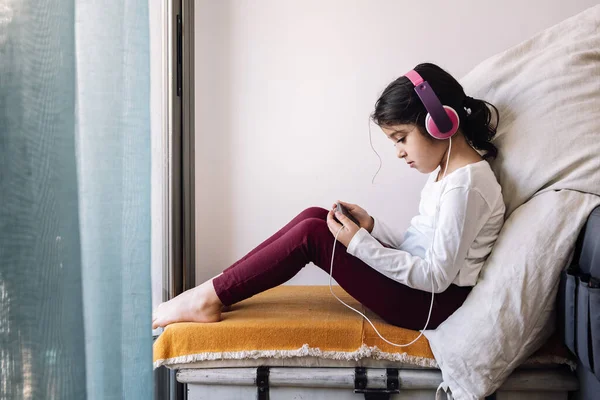little girl sitting in front of her home window with headphones watching videos on the phone, home entertainment for children concept