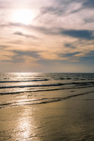 beach with a calm sea and a spectacular sky at sunset, the sun reflects on the water and there is a sailing boat on the horizon, concept of relaxation in nature, vertical photo, copy space for text