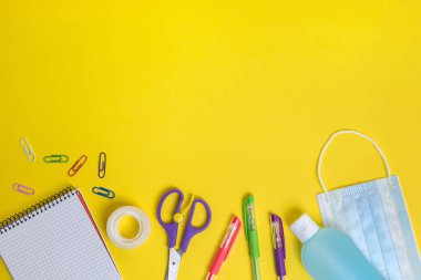 Sarı zemin üzerinde koruyucu maske, dezenfektan jeli ve okul malzemeleri, çocuklar için yeni normallik kavramı ve virüslere karşı koruma, metin için kopyalama alanı