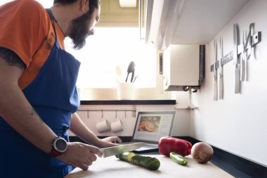 Adam bilgisayarda tarifine danışmanlık yaparken evdeki mutfağın kesme tahtasındaki sebzeleri kesiyor. Sağlıklı yemek kavramı ve yemek yapmayı öğreniyor.