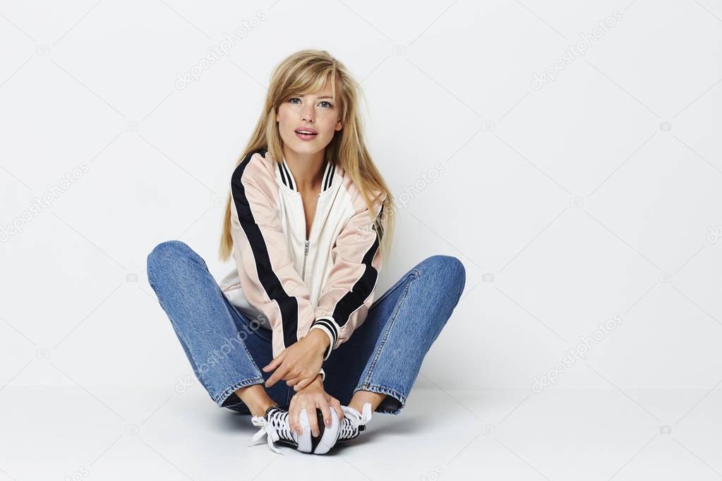 Young woman in jacket and jeans sitting on studio floor and looking at camera