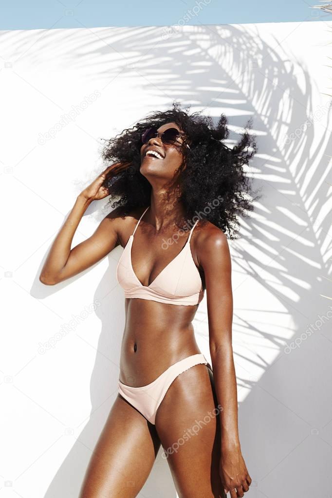 Young african woman in bikini and sunglasses posing at camera standing by white wall