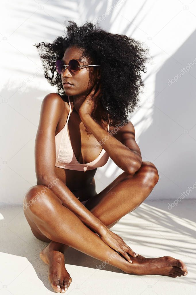 Young african woman in bikini posing at camera sitting by white wall