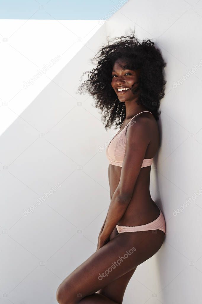 Young african woman in bikini posing at camera leaning by white wall