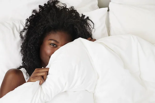 Young african girl peeking over duvet lying in bed, portrait