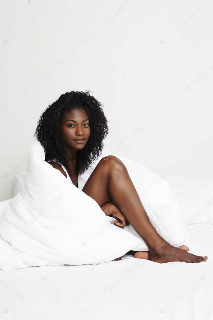 Young african woman sitting in bed wrapped in duvet looking at camera