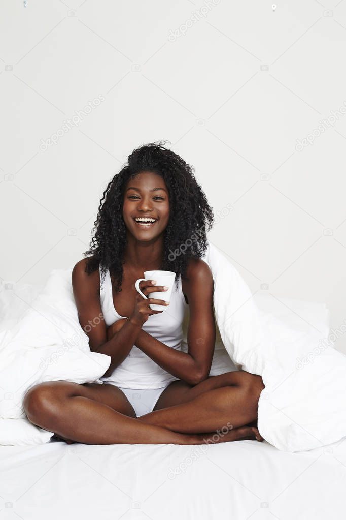 young african woman with coffee cup sitting in bed, smiling at camera