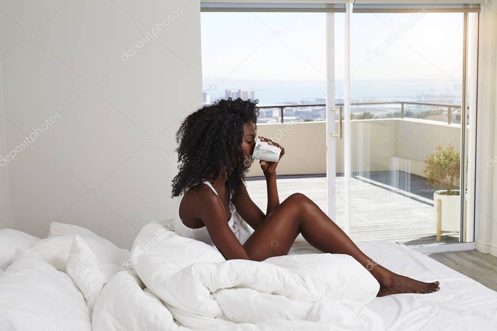 young african woman drinking from coffee cup in bed, side view