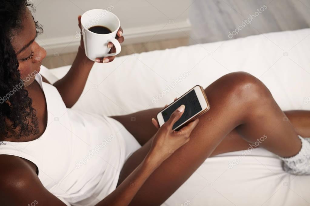 Young african woman in bed with coffee cup texting on smartphone
