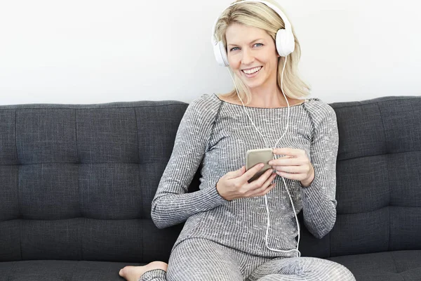 Woman in headphones with smartphone on sofa, smiling at camera