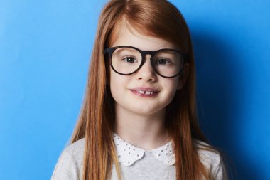 Redheaded girl in spectacles looking at camera, portrait on blue background clipart