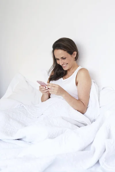中成人黑发妇女坐在床上与智能手机 — 图库照片