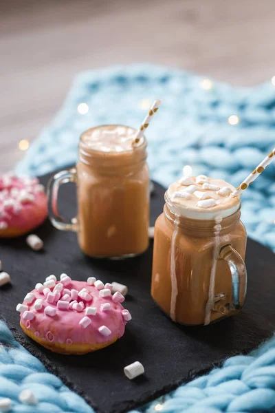 Pink donut with marshmallow and hot chocolate in glass cup on black tray on blue merino knit blanket. Lights on background